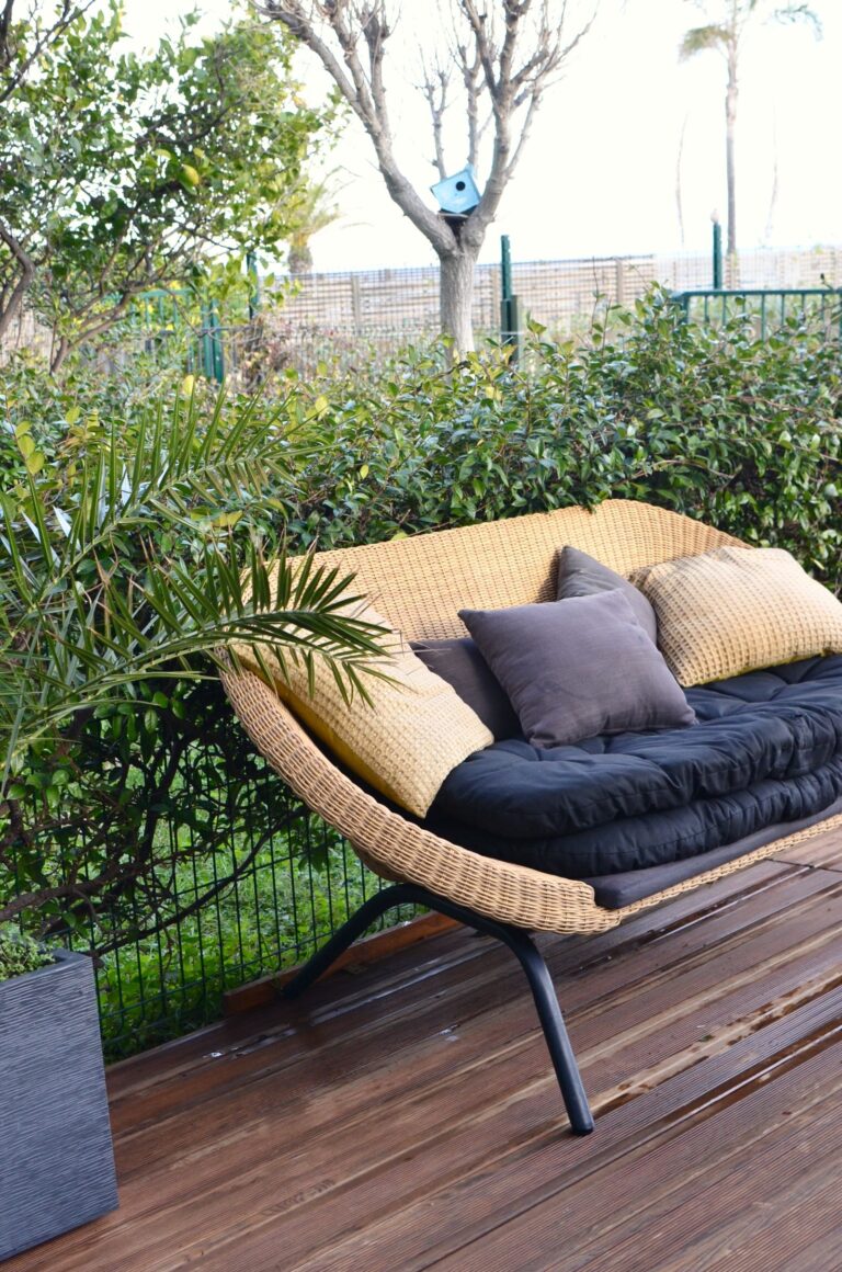 Rieten loungebank met lichte kussens op een houten terras tussen groene planten
